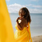 A 50th birthday celebration photoshoot on the coast of Playa del Carmen, showing a woman in a bright yellow dress with an extended train caught in the coastal wind near the ocean waves.