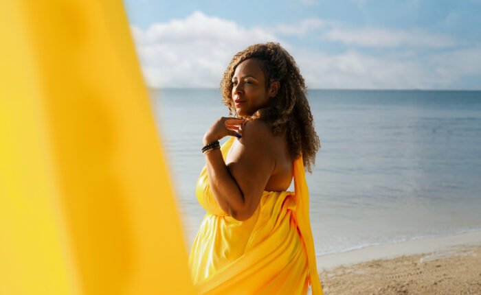 A 50th birthday celebration photoshoot on the coast of Playa del Carmen, showing a woman in a bright yellow dress with an extended train caught in the coastal wind near the ocean waves.