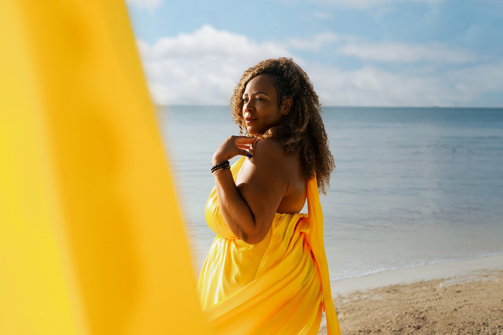 A 50th birthday celebration photoshoot on the coast of Playa del Carmen, showing a woman in a bright yellow dress with an extended train caught in the coastal wind near the ocean waves.